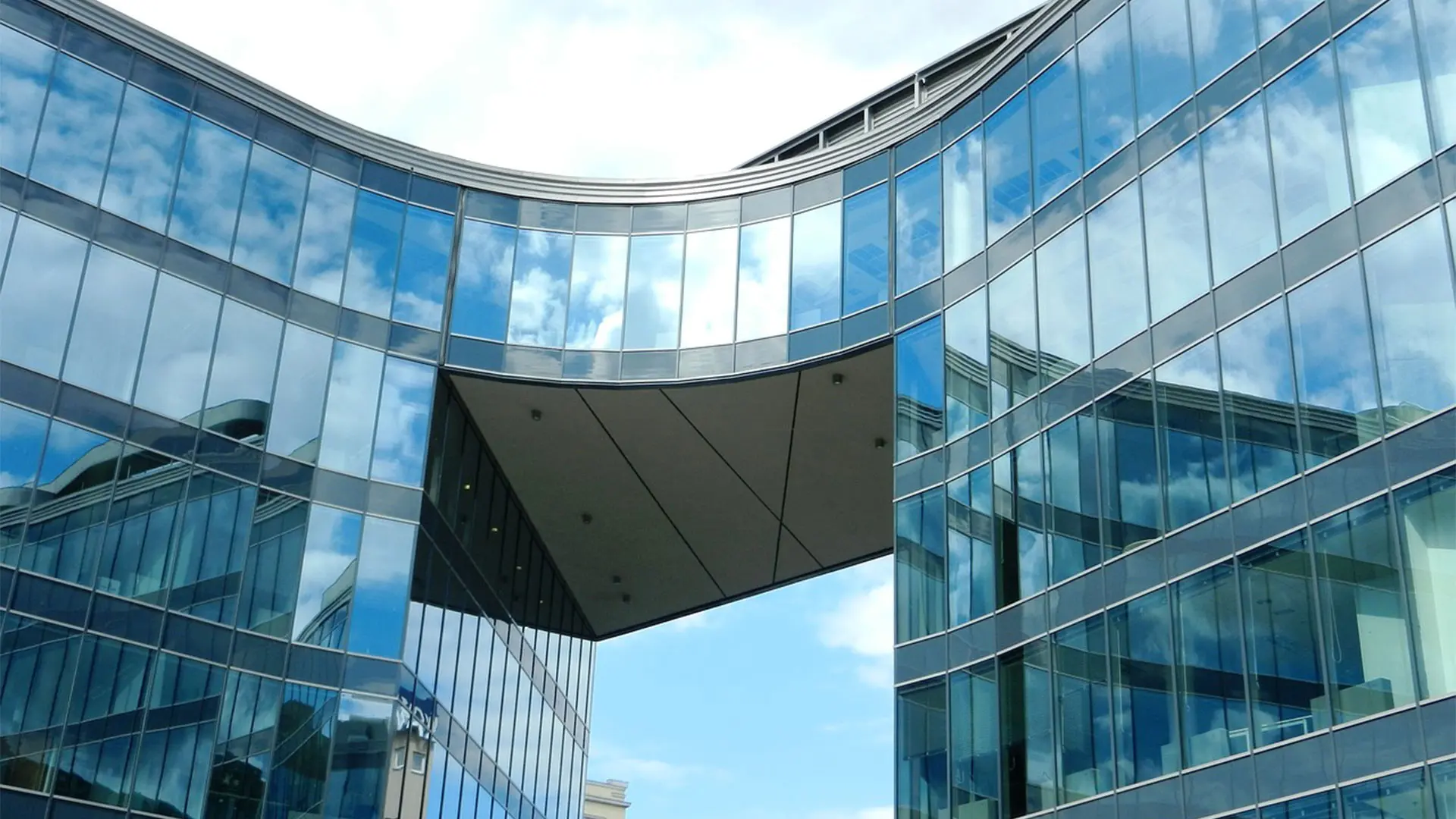 Glass facade of an office building seen from below, reflecting sky and clouds.