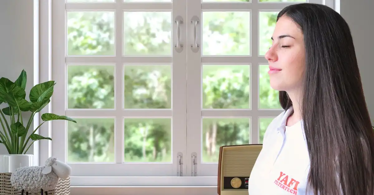 A woman with her eyes closed breathing clean air by an open window, wearing a Yafi Filtertech shirt.